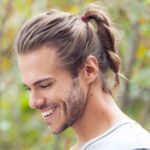 Capture the joyful essence of a man, wearing a classic ponytail hairstyle, as he smiles and gazes downwards, exuding a timeless and confident charm.