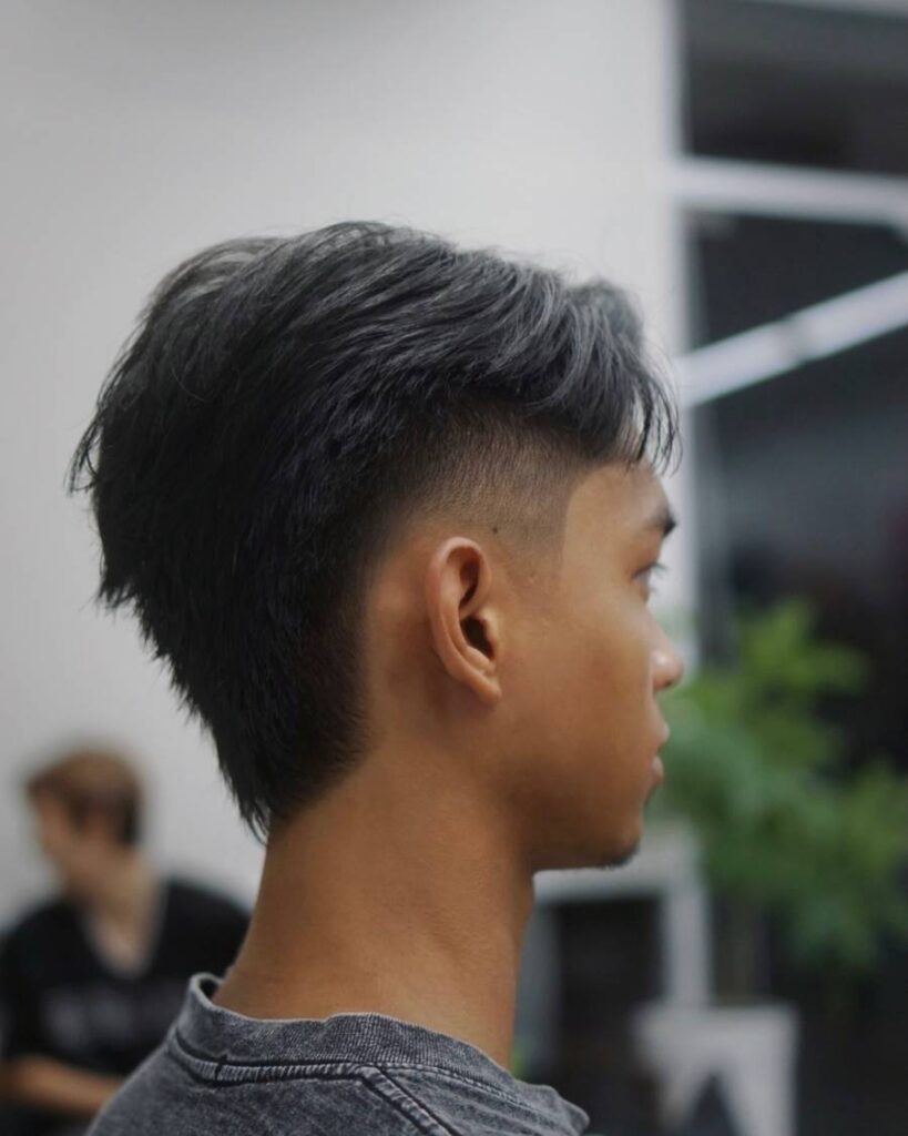 An Asian man is seated with the right side of his head in view, wearing a grey t-shirt. He has black hair styled in a burst fade haircut, emphasizing the sharp fade along the sides.