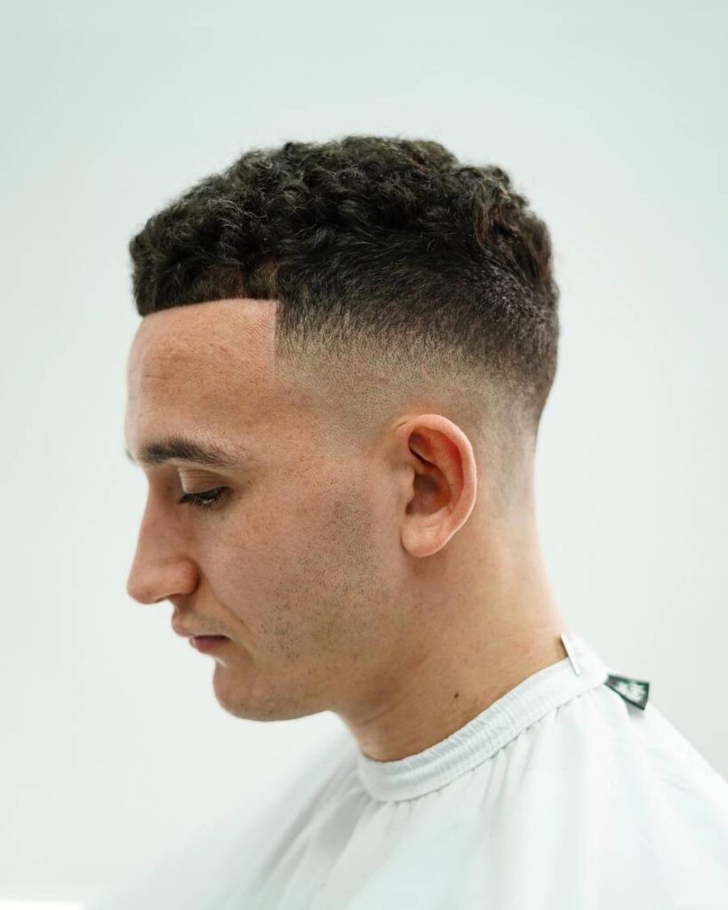 A man with a stylish short drop fade haircut is seated in a barber shop, observing his reflection.